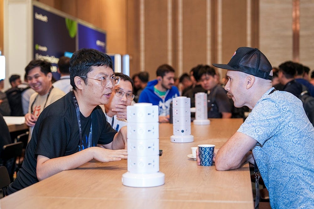 Conference attendees engaged in discussion across a networking table in a busy event hall filled with participants and exhibitor displays.